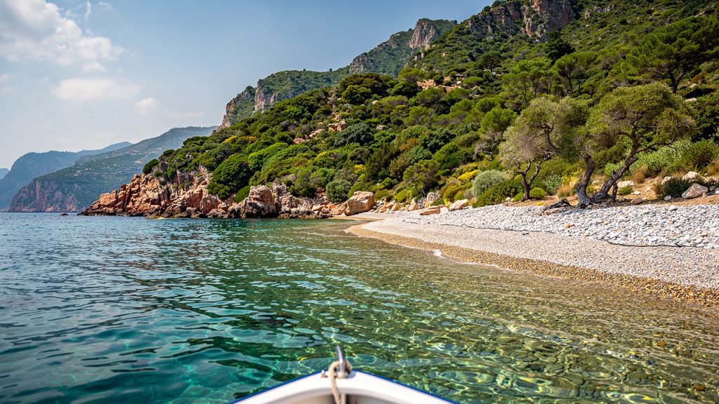 Vue paisible de la baie de Girolata depuis un bateau, avec une eau turquoise limpide, une plage de galets et le maquis corse sous une douce lumière ensoleillée.
